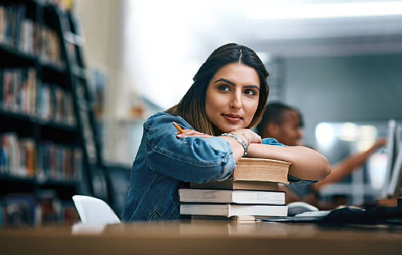 A female graduate student in a library with her arms folded and resting on a stack of text books