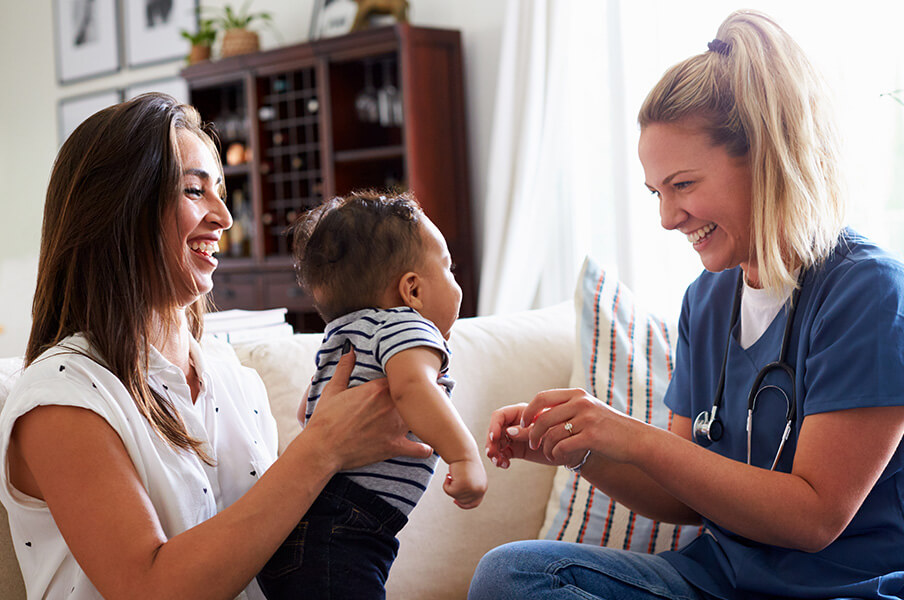 Young mom holding her infant while a smiling visiting nurse looks at child during home visit