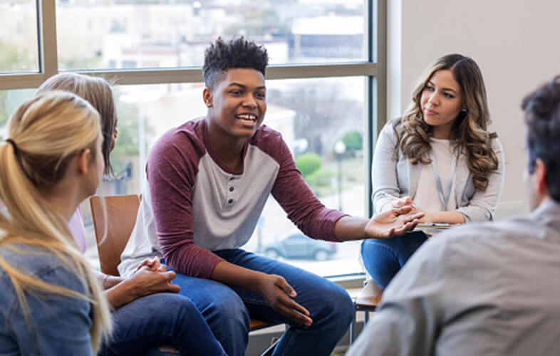 Black teen smiling and speaking in a group therapy session with several participants and a therapist